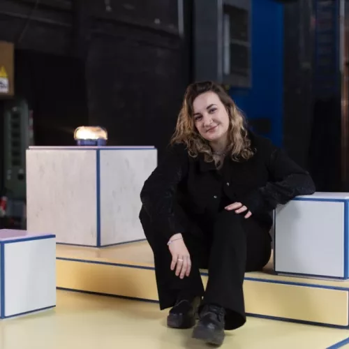 Lotte sitzt auf einem Podest am Set von INTERSECTION. Sie stützt sich auf einen Würfel und lächelt in die Kamera. Sie hat braune, lockige Haare und trägt schwarze Kleidung.