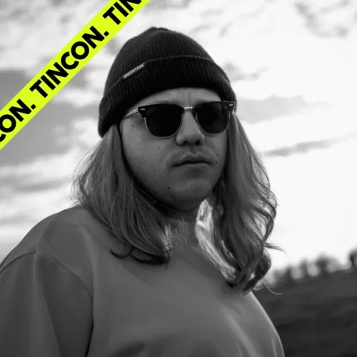 schwarz-weiß Foto von einem Mann mit langen blonden Haaren, er trägt eine Sonnenbrille, eine Mütze und ein lockeres Sweatshirt