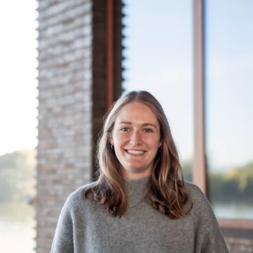 Franziska steht mit grauem Pulli vor Backsteinhaus am Rhein.
