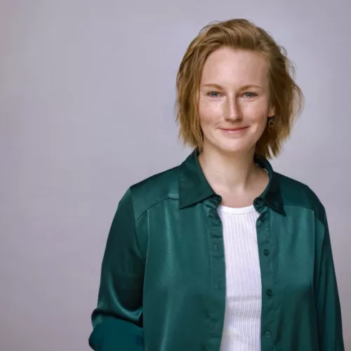 Porträtfoto einer jungen Frau vor hellem, leicht grauen Hintergrund. Die Frau blickt leicht lächelnd in die Kamera, hat kurze, leicht wellige rotblonde Haare und trägt ein dunkelgrünes Hemd über einem hellen Oberteil. Das Bild ist ruhig ausgeleuchtet und zeigt den Oberkörper.