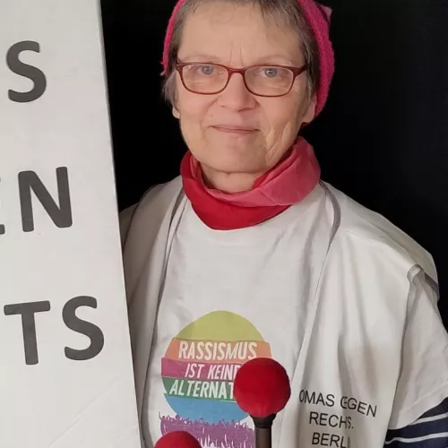 OMA GEGEN RECHTS Franzi mit "Aufstehen gegen Rassismus"-T-Shirt, PussyHat und Trommelschlägeln. Links halb verdeckt A2-Plakat mit Logo der OMAS GEGEN RECHTS