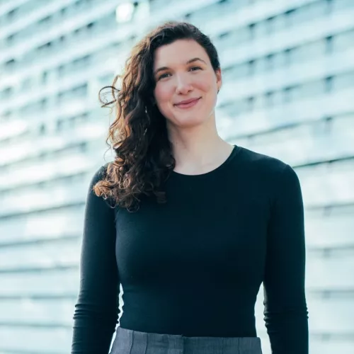 Das Bild zeigt Melanie Weiser im Portraitmodus.  Sie hat lockiges, dunkelbraunes Haar mittellange Länge und trägt ein elegantes, schlichtes, schwarzes Shirt mit langen Ärmeln und graue, faltige Hosen.  Sie steht vor einer Wand mit horizontalen, hellblauen, metallischen Lamellen. 