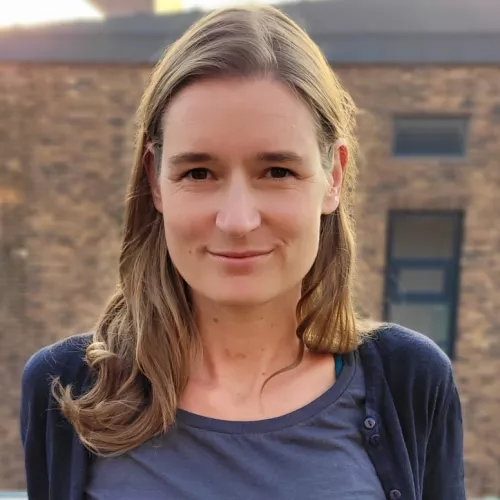 Foto von Kristina Richter im freien vor einer Backsteinfassade im Hintergrund. Offenes Haar, dunkelblaue Kleidung, Lächeln.