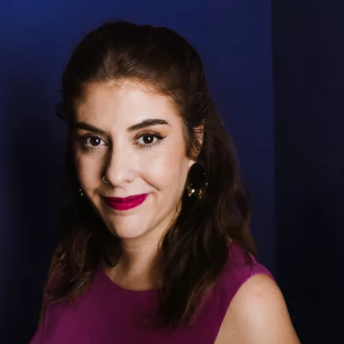 Dr. Milagros Miceli, brown hair and dark eyes, wearing a magenta blouse and lipstick, smiling against a blue wall.