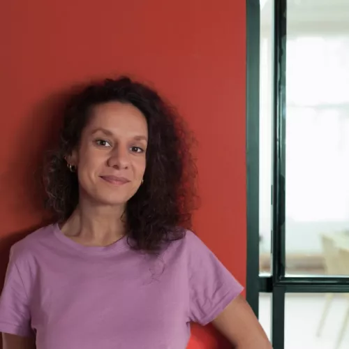 Julia Wadhawan steht vor einer roten Wand, sie trägt ein lila Shirt, die dunklen Locken offen und die Hände hinterm Rücken