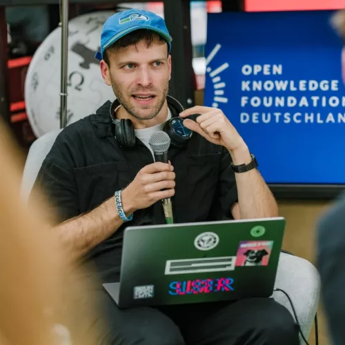Mann Mitte 30 mit blauem Cappy hat einen Laptop auf seinem Schoß und spricht vor Leuten in ein Mikrofon. Im Hintergrund ist ein blaues Schild der Organisation "Open Knowledge Foundation Deutschland" zu sehen.