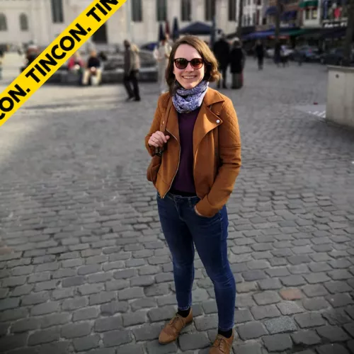 Jana steht auf einem Platz in einer Stadt. Sie trägt Jeans und eine braune Jacke. Sie lächelt in die Kamera