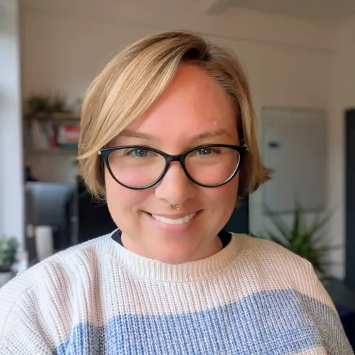 lächelnde Person mit blonden kurzen Haaren und einer schwarzen Brille