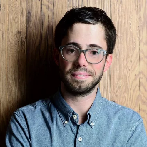 Portrait-Foto eines jungen Mannes vor holzfarbenem Hintergund. Der Mann trägt dunkelbranes Haar, einen Dreitagebart, eine runde Brille und ein blaues Oberhemd.