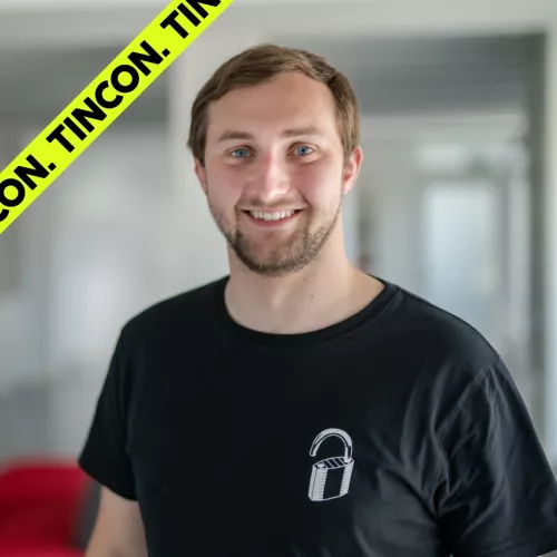 Junger Mann mit kurzen, blonden Haaren und Dreitagebart, trägt ein schwarzes Tshirt und lächelt
