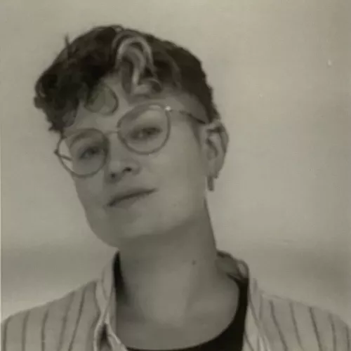 Eine Person mit Brille, gestreiftem Hemd und kurzen Locken schaut mit leicht schiefgelegtem Gesicht in die Kamera.