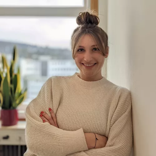 Eine lächelnde Person steht entspannt an eine helle Wand gelehnt und blickt direkt in die Kamera. Die Arme sind locker vor der Brust verschränkt. Die Person hat hellbraunes Haar, das zu einem lockeren Dutt hochgebunden ist, mit einzelnen Strähnen, die das Gesicht einrahmen. Sie trägt einen hellen, grob gestrickten Pullover. Im Hintergrund ist ein Fenster mit unscharfer Aussicht auf Gebäude zu sehen, daneben steht eine Zimmerpflanze auf einem Tisch. Die Atmosphäre wirkt freundlich, ruhig und natürlich.