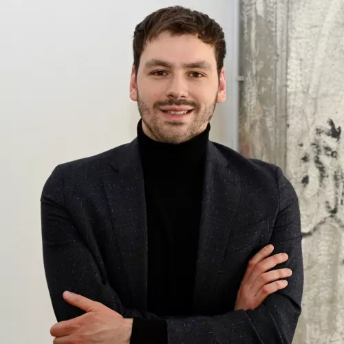 Portrait von Hannes Leiteritz, lächelnd mit verschränkten Armen vor einem weißen Hintergrund mit einer Betonsäule rechts