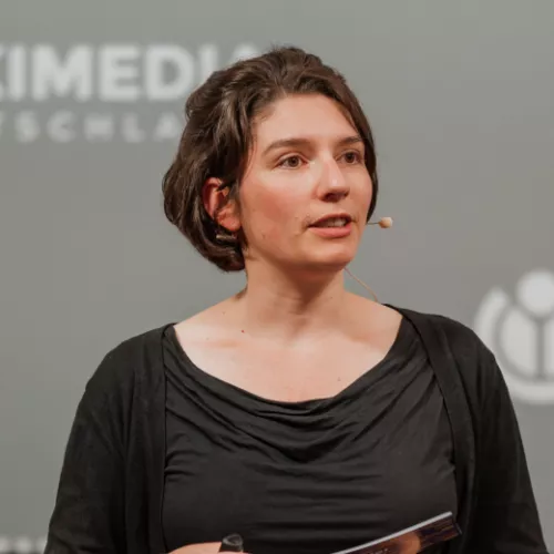 Anne-Sophie Waag während einer Veranstaltung mit Headset und leicht zu Seite blickend ins Publikum. Im Hintergrund eine graue Wikimedia Deustchland Wand mit entsprechendem Schriftzug