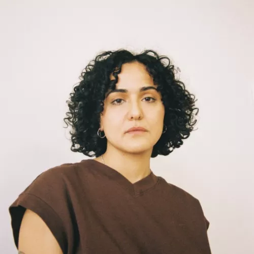 Duygu steht entschlossenen und schaut dezent melancholischen Blickes direkt in die Kamera. Seine*Ihre Schulter ist leicht weggedreht von dem*der Beobachter*in.