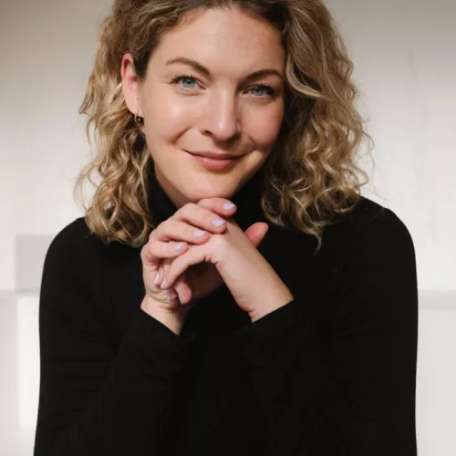 Sally Lisa Starken trägt einen Rollkragenpullover und hat ihr Kinn auf ihren Armen abgestützt. Sie hat Locken und lächelt in die Kamera