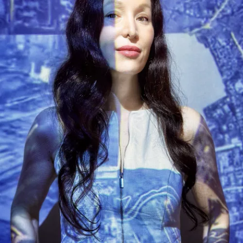 Das Bild zeigt eine weiblich gelesene Person mit langen dunklen Haaren vor einer Wand, auf die eine blaue Computergrafik projiziert wird.