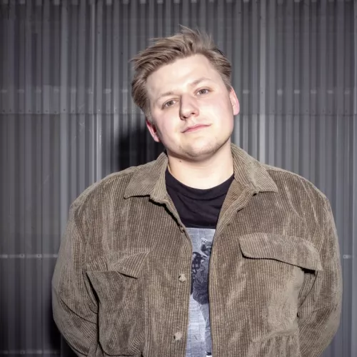 Ein junger Mann in brauner Lederjacke vor dunklem Hintergrund lächelt schief in die Kamera.