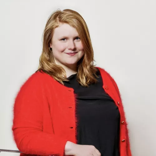 hochkantes Profilfoto: die Dargestellte Person lehnt mit dem rechten Arm auf einer silbernen Stuhllehne und schaut frontal in die Kamera. Sie trägt schwarze Kleidung und eine rote Jacke darüber.