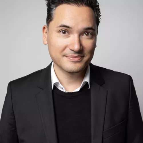 Ein Mann in schwarzem Blazer steht vor einem grauen Hintergrund und lächelt.