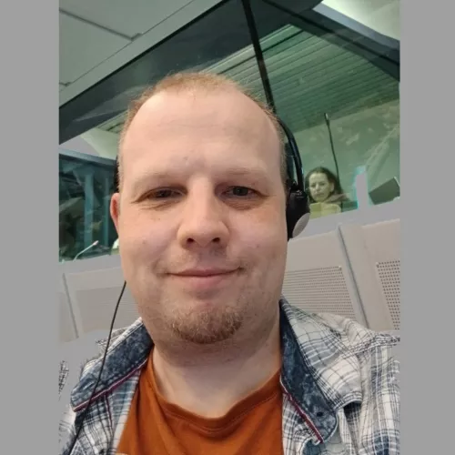 Ein Mann Ende 30 mit kurzgeschorenen blonden Haaren und kurzem Kinnbart. Er trägt ein weißkariertes Hemd über orangenem T-Shirt. Im Hintergrund ist ein Tagungssaal der EU-Kommission zu sehen.
