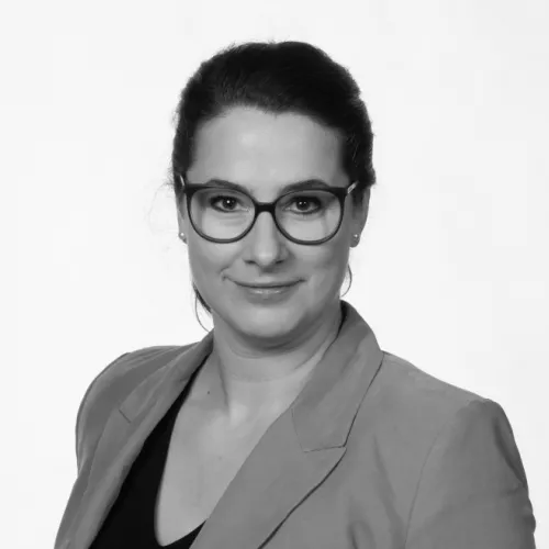 Porträtfoto einer Frau mit dunklen, hochgesteckten Haaren und einer Brille mit dunklem Rahmen. Sie trägt ein helles Sakko über einem dunklen Oberteil. Die Frau lächelt leicht und blickt direkt in die Kamera