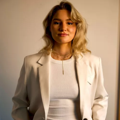Porträt einer Frau mit schulterlangen blonden Haaren vor neutralem Hintergrund. Sie trägt ein helles Oberteil, einen beigen Blazer und eine braune Hose und blickt direkt in die Kamera.