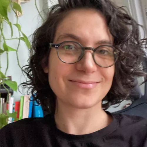 white women, using a black t-shirt and glasses, short-lenght hair, it shows a plant and books at the back