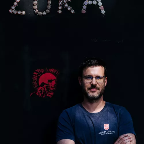 Foto von Robert Fietzke vor einer schwarzen Außenwand in der ZORA. Oben links ist ein Schriftzug aus Kronkorken "ZORA" zu sehen, in der linken Mitte ein rotes Mikro-Graffito mit einem schreienden Punk. Robert Fietzke hat die Arme verschränkt und trägt ein blaues Shirt von der Seenotrettungsorganisation "Mission Lifeline".