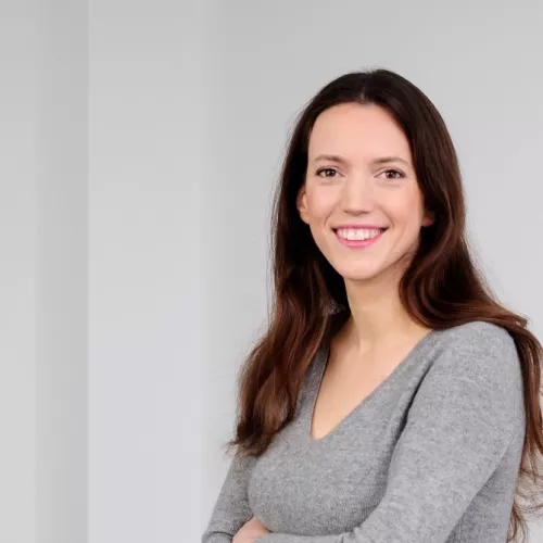 Lena Rohrbach lächelt in die Kamera, sie steht halb seitlich, hat die Arme unter der Brust verschränkt, lange braune Haare und braune Augen und trägt einen hellgrauen Pullover aus Wolle..
