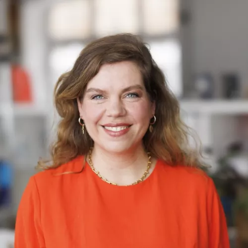 Ein Foto von Jeannette Gusko auf dem sie mit Kopf und Oberkörpr sichtbar ist. Sie trägt ein orangenes Kleid und eine goldene Kette.
