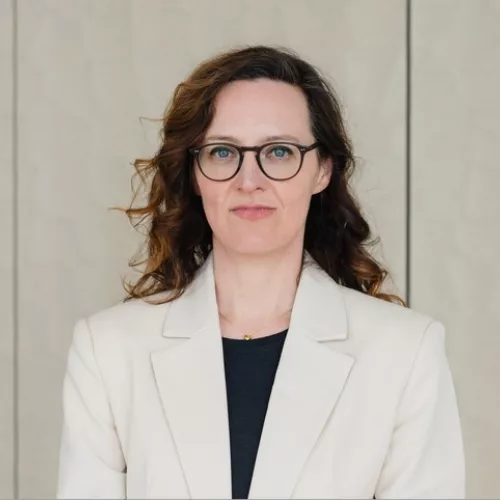 Mittelalte Frau mit langen dunkelbraunen Haaren trägt einen weißen Blazer und ein schwarzes Unterteil.