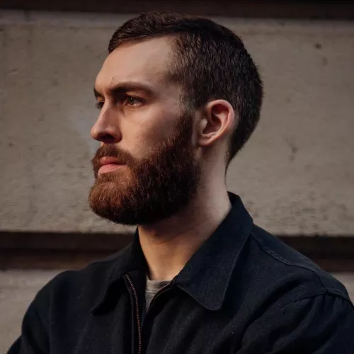 A white man with a red beard looks to the left of the camera.