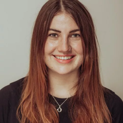 Profilbild von Janne Martha Lentz. Sie ist eine junge, weiße Frau mit langen rot-brauen Haaren. Sie trägt ein schwarzes Shirt und eine silberne Kette.
