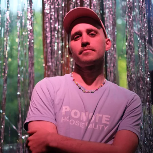 Ich trage ein pinkes Cappy und ein hellblaues Shirt, stehe vor einem Glitzervorhang in pink angehauchtem Licht.