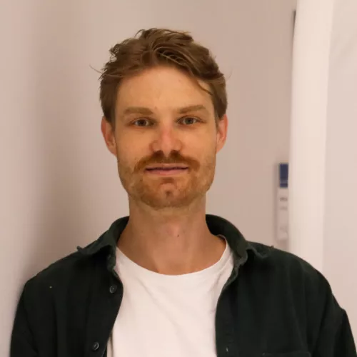 Ein junger Mann blickt direkt in die Kamera. Er hat kurzes, leicht zerzaustes blondes Haar und einen braun-blonden Schnurrbart mit kurzem Bart am Kinn. Er trägt ein weißes T-Shirt unter einem dunklen offenen Hemd. Der Hintergrund ist hell und unscharf und sieht aus wie ein Innenraum mit weißen Wänden.