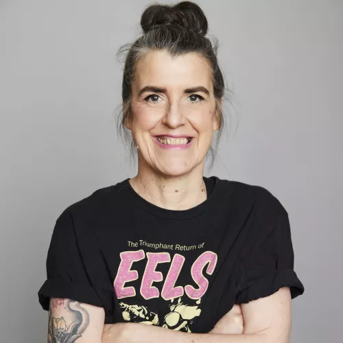 Eine weibliche Person mit verschränkten Armen lächelt ins Bild, auf ihrem T-Shirt ist der Bandname EELS zu lesen.