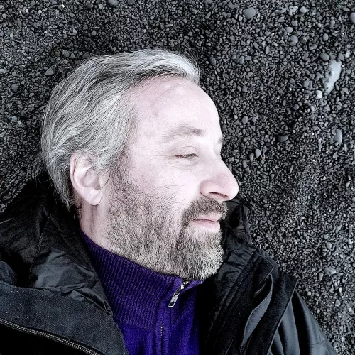 Picture of Robin Berjon, white skin, silver hair, beard, looking right, on a black volcanic sand background