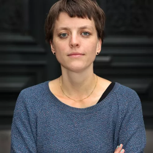 Sarah Reimann steht vor einer schwarzen Tür. Sie ist weiß, hat kurze Haare und trägt einen blauen Pullover.