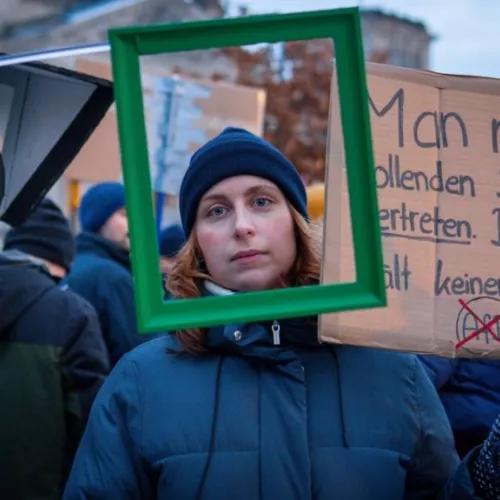 Frau mit grünen Rahmen um das Gesicht, im Hintergrund links ein großer Kamerakopf und rechts ein Demoschild.