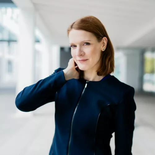 Foto von Vivian Pein, eine rothaarige Frau in einem blauen Blazer, die lächelnd vor einem Gebäude steht