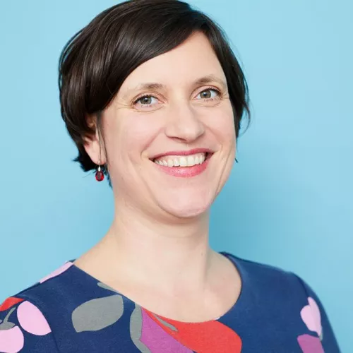 Maite Woköck schaut lächelnd direkt in die Kamera. Sie trägt kurze Haare, ein geblümtes Oberteil und steht vor einem hellblauen Hintergrund. 